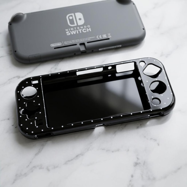 May include: A dark gray Nintendo Switch Lite console positioned above a black protective case featuring a white polka dot pattern. The console displays the Nintendo Switch logo on its back, while the case has cutouts for buttons and the screen.