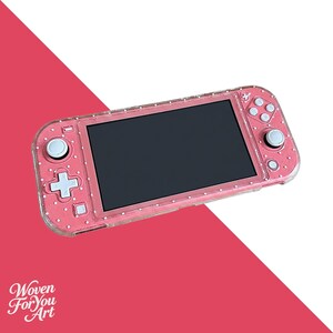 May include: A handheld gaming console featuring a pink shell with a white polka dot pattern and a clear protective case. The device includes a black screen, white control buttons, and two analog sticks. The text Woven For You Art is visible.