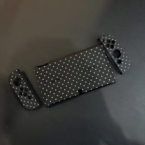 May include: A black gaming console and two detachable controllers, all featuring a white polka dot pattern. The console is rectangular with a central screen area and a kickstand, while the two controllers have circular button cutouts.