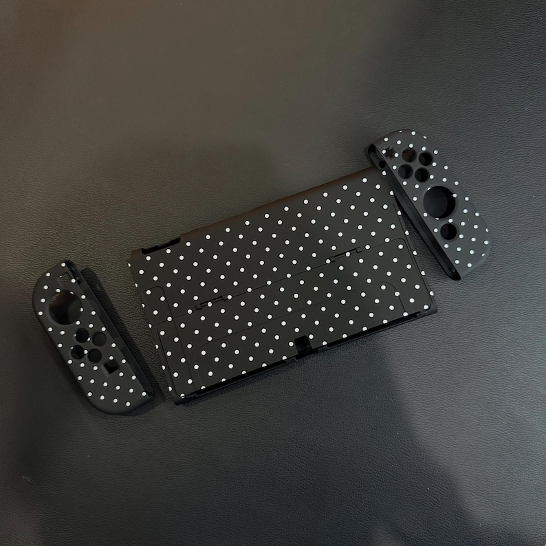 May include: A black gaming console and two detachable controllers, all featuring a white polka dot pattern. The console is rectangular with a central screen area and a kickstand, while the two controllers have circular button cutouts.