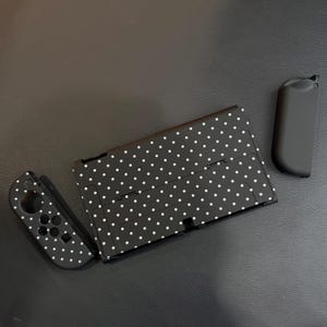 May include: A black gaming console back plate and one matching controller attachment, both featuring a white polka dot pattern. A separate solid black controller attachment is positioned to the right of the console component.