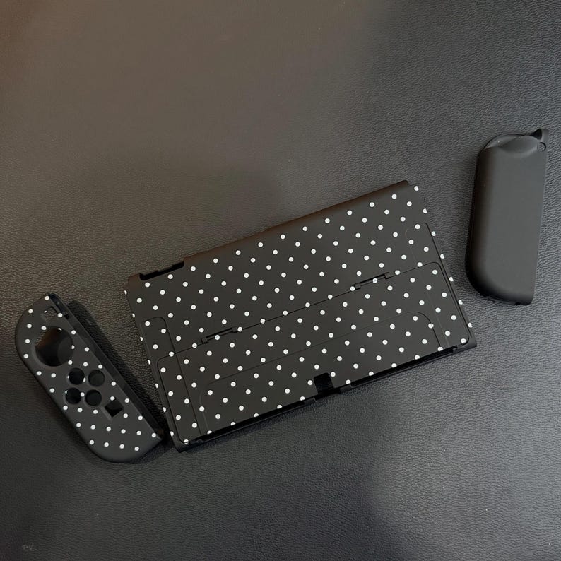 May include: A black gaming console back plate and one matching controller attachment, both featuring a white polka dot pattern. A separate solid black controller attachment is positioned to the right of the console component.