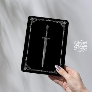 May include: A black rectangular product featuring a silver sword design. The sword is centered on the product, with a decorative border. The product is held by a hand, with the text "Woven For You Art" in the lower right corner.