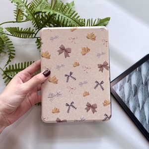 May include: A beige rectangular case featuring a pattern of brown bows and autumn leaves. The case, held in a hand, is likely for an e-reader or tablet. The design is repeated across the surface.
