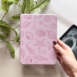 Puede incluir: Una funda rosa para lector de libros electrónicos con un patrón repetido de conchas marinas, estrellas de mar y burbujas en un tono rosa más oscuro. La funda es rectangular con esquinas redondeadas. Una mano con esmalte de uñas rojo oscuro sostiene la funda.