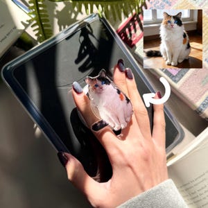 May include: A clear phone grip featuring a calico cat with blue eyes. The cat image is on a clear, circular grip, attached to a black phone case. The grip is held up by a hand with dark nail polish. A photo of the cat is in the background.
