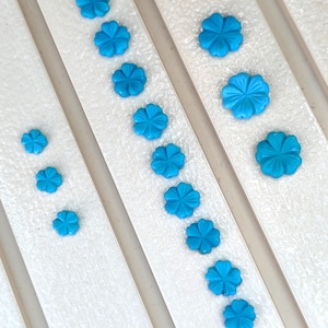 May include: A collection of bright turquoise blue flower-shaped cabochons arranged in rows. The set includes various sizes of five-petaled floral pieces with a smooth, glossy finish, displayed in a linear formation on a white textured surface.