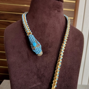 Puede incluir: Un collar de serpiente en tono dorado con una cabeza incrustada de pequeñas cuentas turquesa, una piedra turquesa ovalada central y detalles rojos. El cuerpo flexible se compone de eslabones dorados entrelazados con filas de cuentas turquesa.