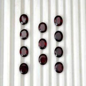 Pode incluir: Um conjunto de nove pedras preciosas granada vermelho-escuras, lapidadas em formato oval e dispostas em três colunas verticais. Cada gema apresenta uma superfície facetada que reflete a luz, exibindo um tom carmesim profundo e uma forma oval polida.