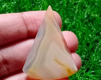 May include: A triangular-shaped, polished gemstone with a layered pattern of beige, white, and light orange hues. The stone is held in a hand, with a green grassy background.