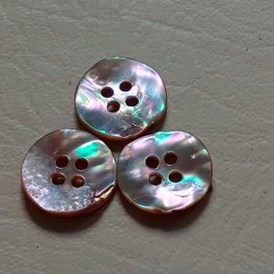 Puede incluir: Tres botones redondos iridiscentes, cada uno con cuatro agujeros. Los botones tienen un brillo nacarado con tonos rosas, verdes y azules. Los botones están dispuestos en un patrón triangular.