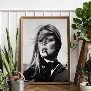 May include: Monochrome framed print of a woman smoking, with her hair blowing in the wind. The image is in a wooden frame, with plants and a watering can in the foreground.