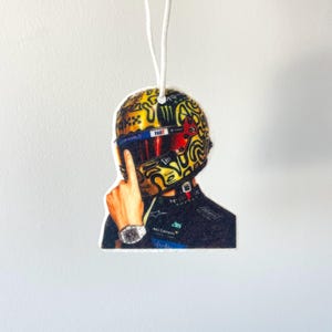 Peut inclure: Désodorisant pour voiture représentant un casque de course avec un motif or et noir. Le casque a une visière rouge et le mot "FART" est visible. Le désodorisant est suspendu par une ficelle blanche.