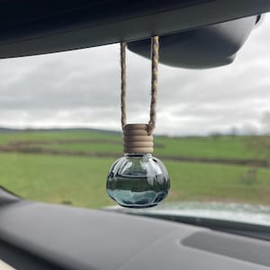 Op de afbeelding: Een heldere, ronde glazen auto luchtverfrisser met een lichtblauwe vloeistof erin, opgehangen aan een bruin touw. De houten dop zit bovenop de glazen bol. De achtergrond toont een groen veld en een bewolkte lucht.