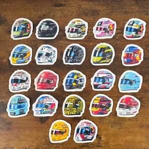 May include: A collection of twenty-two individual stickers featuring various Formula One racing helmet designs. Each sticker displays a unique colour scheme, sponsor logos, and visor details, arranged in a grid pattern on a wooden surface.