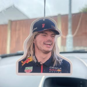 May include: Car air freshener with an image of a smiling man in a navy racing uniform and a number 1 baseball cap. The freshener has a hanging string and features the Oracle Red Bull Racing and Tag Heuer logos.