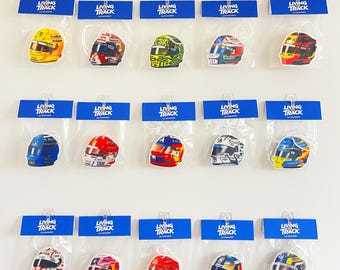 May include: Fifteen individual air fresheners shaped like racing helmets, each packaged in a clear plastic sleeve with a blue Living Track header card. The helmets feature various colorful designs, logos, and patterns representing different racing teams.