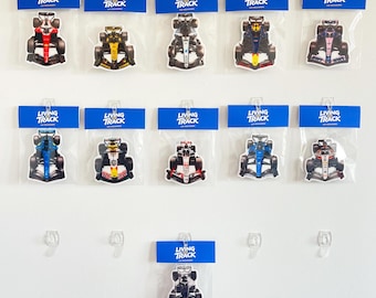 May include: Eleven individual Formula 1 car air fresheners, each packaged in a clear plastic sleeve with a blue header card labeled Living The Track. The collection features various racing car designs in distinct color schemes and liveries.