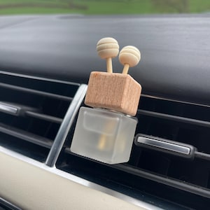 Puede incluir: Un ambientador para coche con un frasco de vidrio esmerilado y una parte superior de madera con dos detalles esféricos. El ambientador está montado en una rejilla de ventilación del coche, con un salpicadero de color claro y un campo verde al fondo.