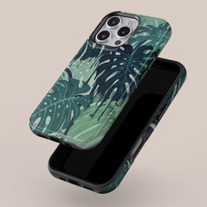 May include: A smartphone case with a tropical leaf design in shades of green and dark blue. The case is shown detached from a black smartphone, revealing the phone's screen. The design features large monstera leaves.