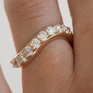 Puede incluir: Un anillo de oro curvado engastado con múltiples piedras preciosas pequeñas y claras. El anillo se lleva en un dedo, mostrando su elegante diseño y piedras brillantes. La banda del anillo se curva suavemente.