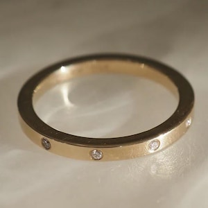 May include: A gold-colored ring with small, evenly spaced clear gemstones. The ring is circular and sits on a white surface. The ring is a simple band design, suitable for everyday wear.