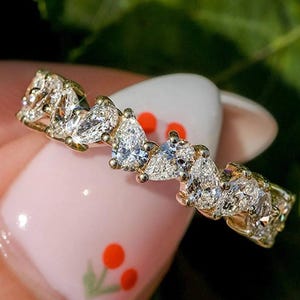 May include: A gold ring with a series of pear-shaped diamonds. The ring is set with multiple clear gemstones, creating a sparkling effect. The ring is held against a background of green leaves.