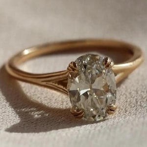Puede incluir: Un anillo de oro con un diamante de talla ovalada. El anillo tiene una banda delicada y un engaste de cuatro garras. El diamante es claro y refleja la luz. El anillo está sobre un fondo suave y neutro. Diseño elegante.