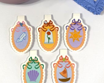 Puede incluir: Cinco imanes decorativos de forma ovalada con ilustraciones de cuentos de hadas. Cada imán presenta un diseño diferente: un zapato de cristal, una rosa en un vaso, un sol, una concha y un velero. Los imanes tienen un fondo blanco y están bordeados en naranja.