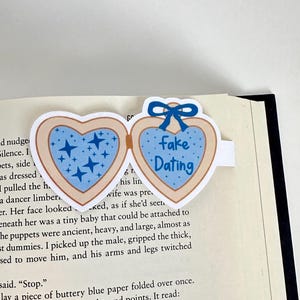 May include: A heart-shaped bookmark with a blue bow and the words "Fake Dating." The bookmark has two heart-shaped sections, one with blue stars and the other with the text. The bookmark is on a book page.
