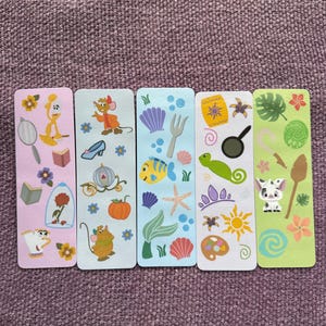 May include: Five colorful rectangular bookmarks featuring Disney characters and themed illustrations. Each bookmark has a different background color and depicts characters and objects from various Disney movies, including Cinderella, Beauty and the Beast, and The Little Mermaid.