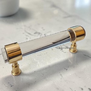 May include: A cabinet pull featuring a clear, cylindrical acrylic center bar accented by polished gold-tone metal end caps and mounting posts. The hardware includes the small text home.crafthardware printed on the side of the clear acrylic bar.