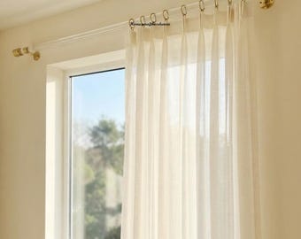 May include: A clear acrylic curtain rod with gold-toned metal brackets and end caps, featuring gold-toned rings holding a sheer off-white curtain. The text Homecrafthardware is printed in black on the curtain fabric near the top rings.