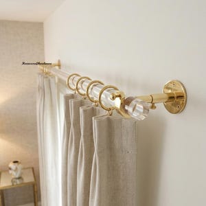 May include: A clear acrylic curtain rod with polished gold metal brackets and end caps. The rod features gold metal rings holding beige fabric curtains. The text Homexcrafthardware is visible on the wall near the left mounting bracket.