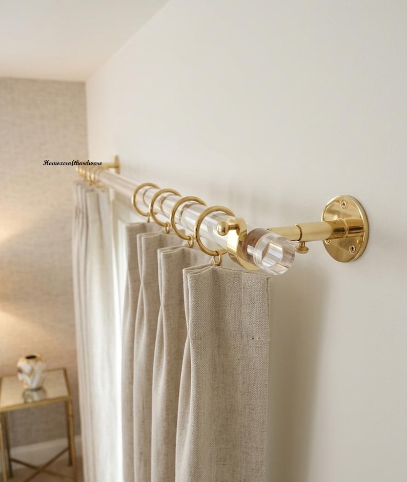 May include: A clear acrylic curtain rod with polished gold metal brackets and end caps. The rod features gold metal rings holding beige fabric curtains. The text Homexcrafthardware is visible on the wall near the left mounting bracket.