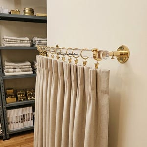 May include: A clear acrylic curtain rod with polished gold-tone metal wall brackets and end caps. The rod features a series of gold-tone metal rings with attached clips, holding a pleated beige fabric curtain. The text Homecrafthardwa is visible.