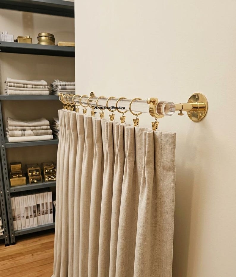 May include: A clear acrylic curtain rod with polished gold-tone metal wall brackets and end caps. The rod features a series of gold-tone metal rings with attached clips, holding a pleated beige fabric curtain. The text Homecrafthardwa is visible.