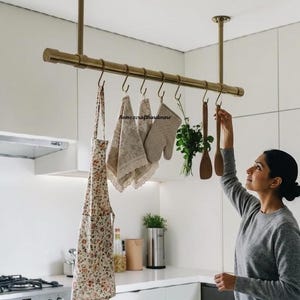 Könnte beinhalten: Ein an der Decke montierter Küchen-Topfhalter aus Messing mit einer horizontalen Stange, die von zwei vertikalen Stützen gehalten wird. Sieben S-förmige Haken hängen an der Stange und tragen eine geblümte Schürze, zwei Spitzenstoffe, einen beigen Ofenhandschuh, Kräuter und Holzlöffel.