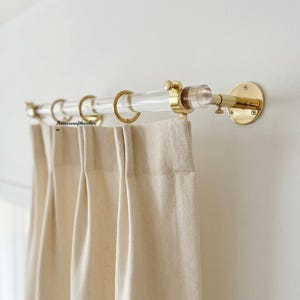 May include: A clear acrylic curtain rod with polished brass mounting brackets and matching brass rings. The rod supports beige, pleated fabric curtains. The text "Homexcrafthardware" is visible on the left side of the curtain rod assembly.
