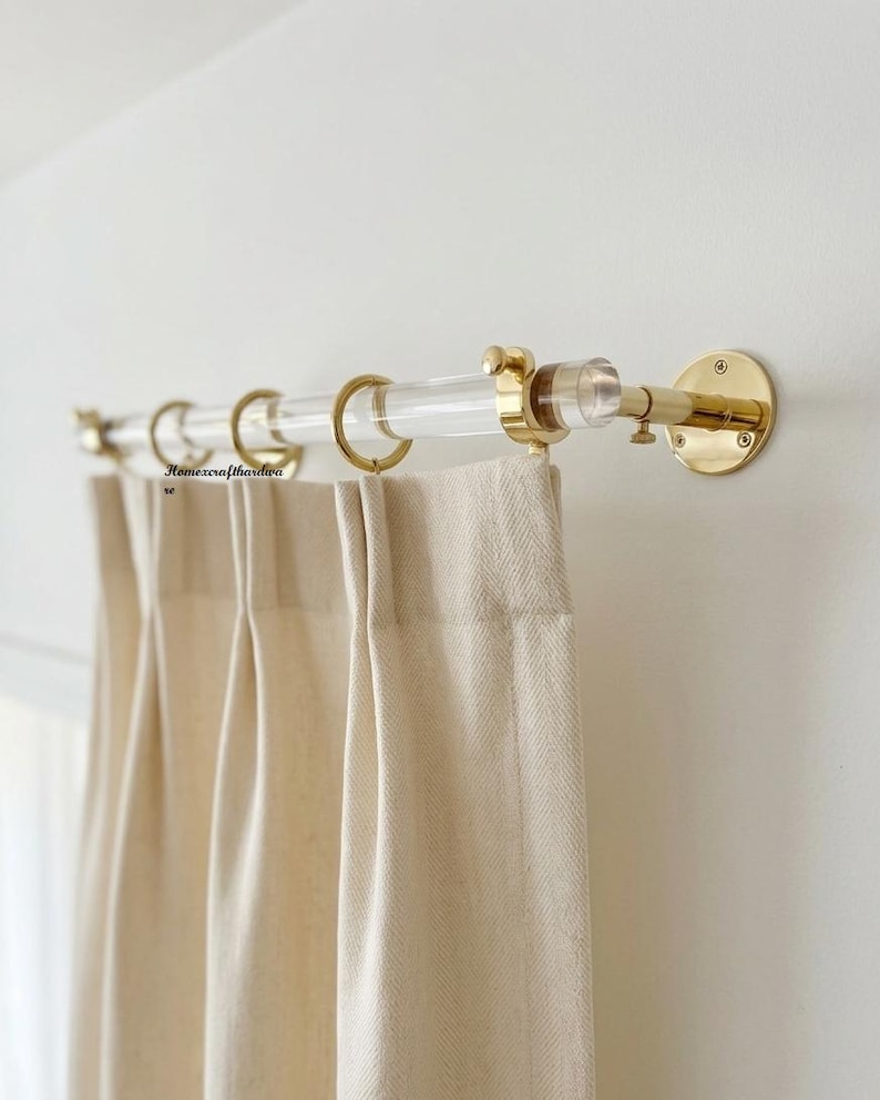May include: A clear acrylic curtain rod with polished brass mounting brackets and matching brass rings. The rod supports beige, pleated fabric curtains. The text "Homexcrafthardware" is visible on the left side of the curtain rod assembly.