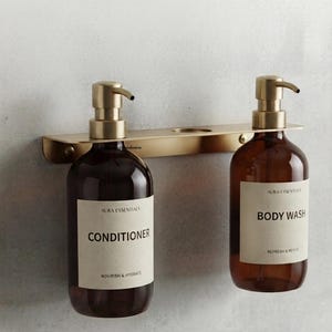 Op de afbeelding: Een aan de muur gemonteerd rek van goudkleurig metaal met twee amberkleurige glazen flessen met pompdoseerders. De linkerfles heeft het label Aura Essentials Conditioner en de rechterfles Aura Essentials Body Wash, beide met zwarte tekst.