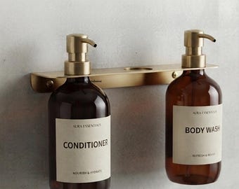 May include: A wall-mounted gold metal rack holding two amber glass bottles with pump dispensers. The left bottle is labeled Aura Essentials Conditioner, and the right bottle is labeled Aura Essentials Body Wash, both featuring black text.