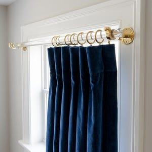 May include: A clear acrylic curtain rod with polished gold-tone metal brackets and end caps, featuring gold-tone metal rings holding a deep navy blue velvet curtain panel. The rod is mounted above a window frame with visible gold-tone hardware.