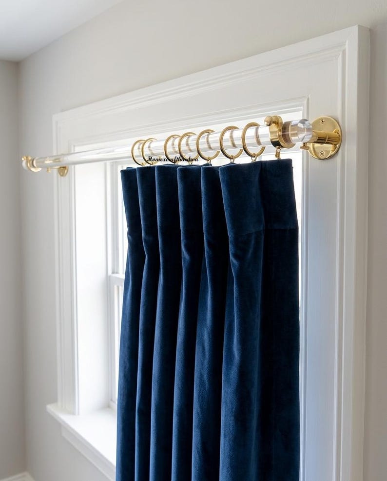 May include: A clear acrylic curtain rod with polished gold-tone metal brackets and end caps, featuring gold-tone metal rings holding a deep navy blue velvet curtain panel. The rod is mounted above a window frame with visible gold-tone hardware.