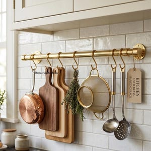 May include: A polished brass kitchen hanging rack mounted on a white tiled wall. The rack features S-hooks holding a copper pan, two wooden cutting boards, a bundle of herbs, a gold mesh strainer, a ladle, a slotted spoon, and a tag reading KITCHEN USED BRASS HANGING RACK.