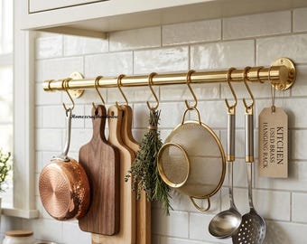 May include: A polished brass kitchen hanging rack mounted on a white tiled wall. The rack features S-hooks holding a copper pan, two wooden cutting boards, a bundle of herbs, a gold mesh strainer, a ladle, a slotted spoon, and a tag reading KITCHEN USED BRASS HANGING RACK.