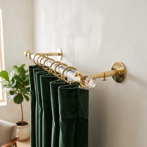 May include: A curtain rod featuring a clear acrylic pole with polished gold-tone metal wall brackets and matching gold-tone rings. The rod is mounted horizontally, holding dark green velvet curtains that hang in vertical folds from the rings.