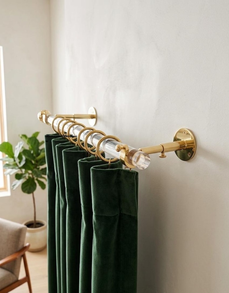 May include: A curtain rod featuring a clear acrylic pole with polished gold-tone metal wall brackets and matching gold-tone rings. The rod is mounted horizontally, holding dark green velvet curtains that hang in vertical folds from the rings.