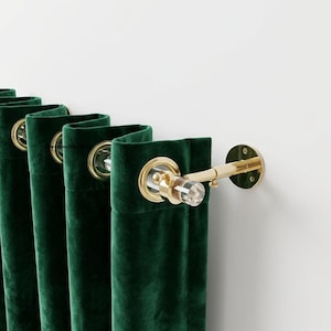 May include: Deep emerald green velvet curtains featuring gold-toned metal grommets. The curtains are suspended on a clear acrylic rod with gold-toned metal wall brackets and a matching clear acrylic finial at the end of the curtain rod.