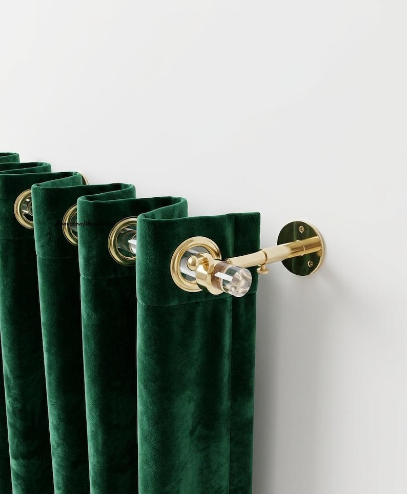 May include: Deep emerald green velvet curtains featuring gold-toned metal grommets. The curtains are suspended on a clear acrylic rod with gold-toned metal wall brackets and a matching clear acrylic finial at the end of the curtain rod.